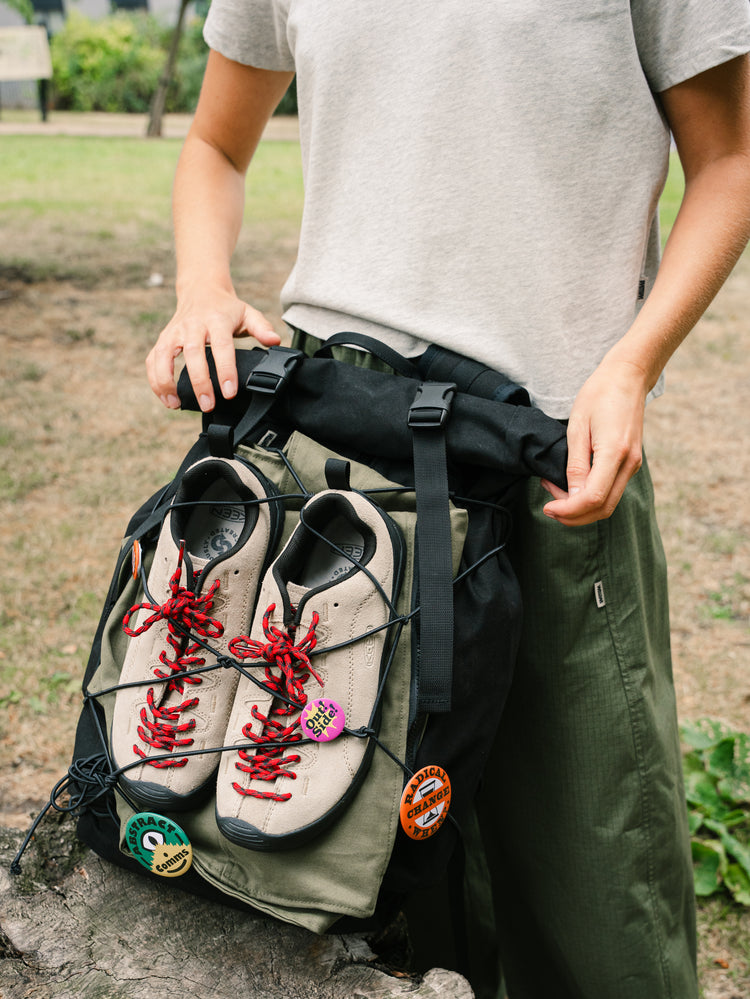 Bridge Backpack in Black outdoors with shoes secured under the front bungee cord